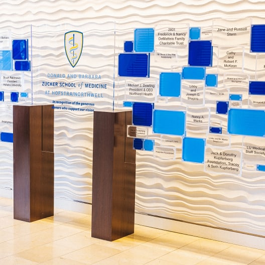 A donor recognition wall at the Zucker School of Medicine, featuring blue and clear plaques with donor names, mounted on a wavy white background with wooden supports.