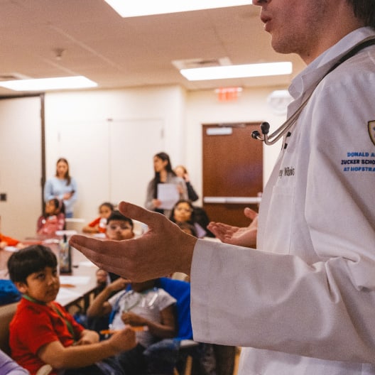 A doctor in a white coat speaks to a group of children seated at tables in a classroom, while a few adults observe in the background. The focus is on the doctor’s hand and the children’s attentive faces.