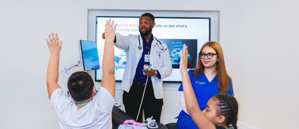 A male doctor in a white coat and a female nurse in blue scrubs interact with children in a classroom; one child raises their hand while the doctor gestures and smiles. A white screen with text and images is in the background.