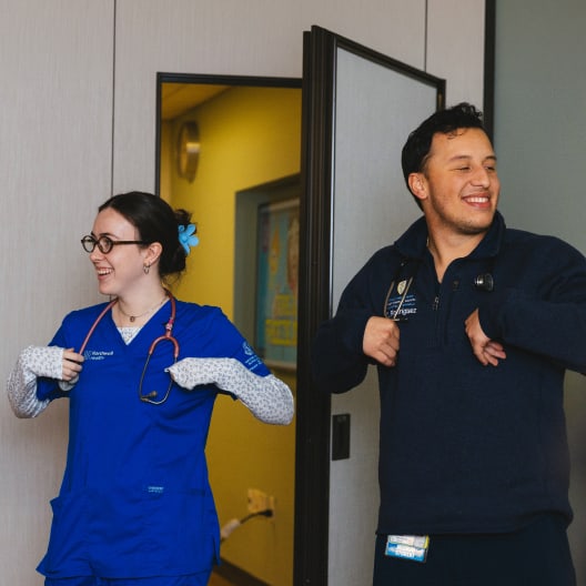 Two healthcare workers smiling and standing indoors. One wears blue scrubs with a stethoscope, glasses, and a blue hair clip; the other wears a navy pullover and name badge. Both have their hands at their chests, appearing cheerful.