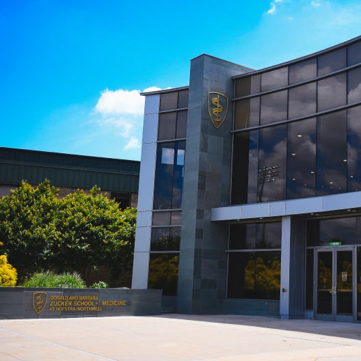 Entrance of the Zucker School of Medicine at Hofstra/Northwell, featuring a modern glass building with a medical insignia, greenery in the background, and clear blue sky overhead.
