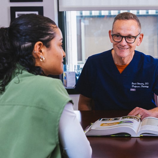 A woman sits across a desk from a smiling male doctor in scrubs, having a conversation. An open book is on the desk between them, and the setting appears to be a medical office.