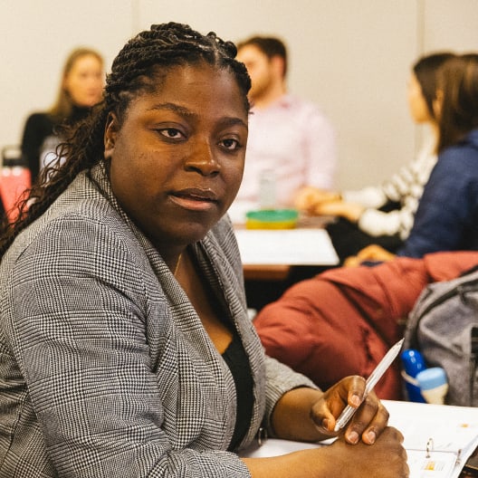A woman in a gray blazer sits at a table holding a pen, looking at the camera. In the background, several people are engaged in conversation in a classroom or meeting setting.