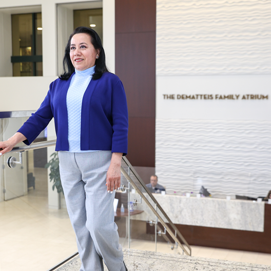 A woman with dark hair, wearing a blue jacket and light blue top, stands on a staircase in a modern atrium. The sign behind her reads “The Dematteis Family Atrium.”.