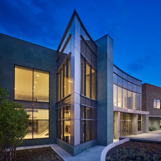 Modern building with large glass windows and a well-lit interior, photographed at dusk. The exterior features clean lines, sharp angles, and a combination of glass and stone surfaces against a deep blue evening sky.