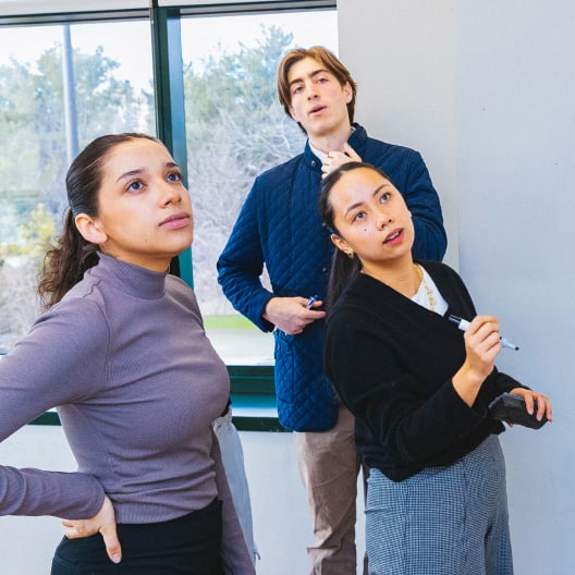 Three people stand indoors near a window, looking upward thoughtfully. One woman holds a marker as if explaining something on a board, while the others listen and observe attentively. Trees are visible outside.