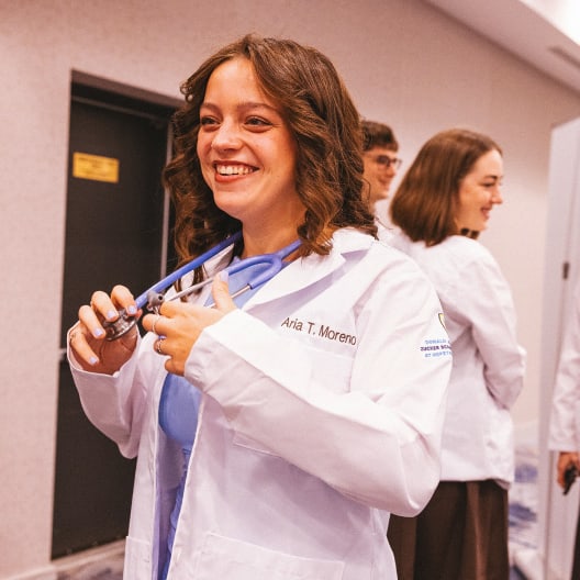 A smiling woman in a white lab coat with a stethoscope around her neck stands in a hallway with other people in similar coats, suggesting a medical or healthcare setting.