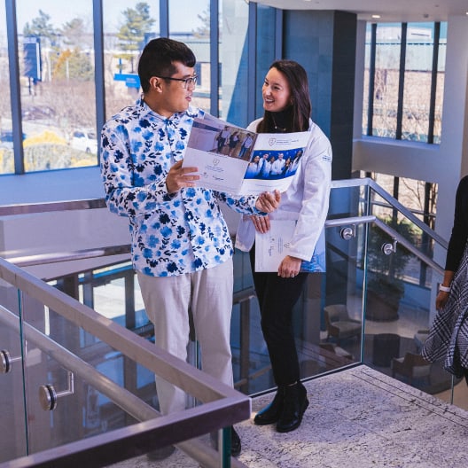 A man and a woman stand on a staircase indoors, smiling and holding papers. The woman wears a white coat and the man wears a blue and white patterned shirt. They appear to be talking in a brightly lit building.