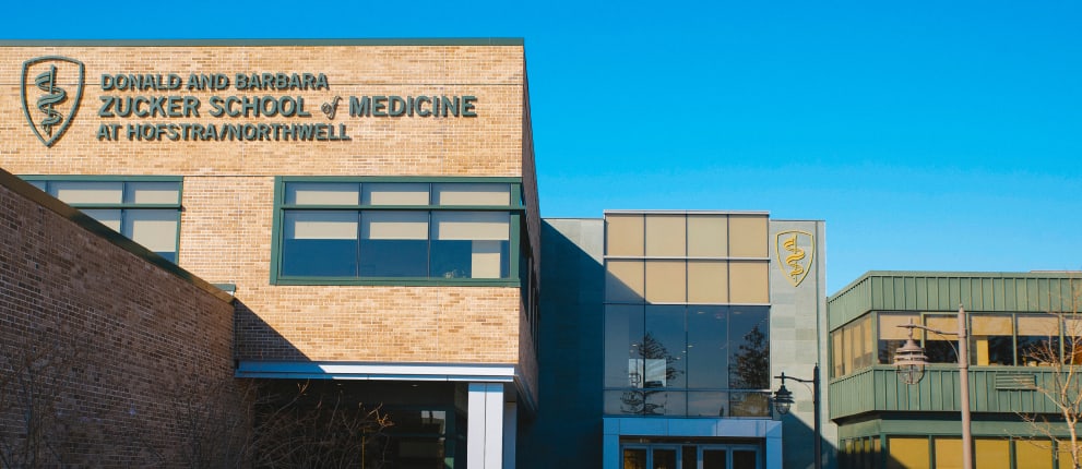 The exterior of the Donald and Barbara Zucker School of Medicine at Hofstra/Northwell, featuring large building signs and a medical caduceus symbol, under a clear blue sky.