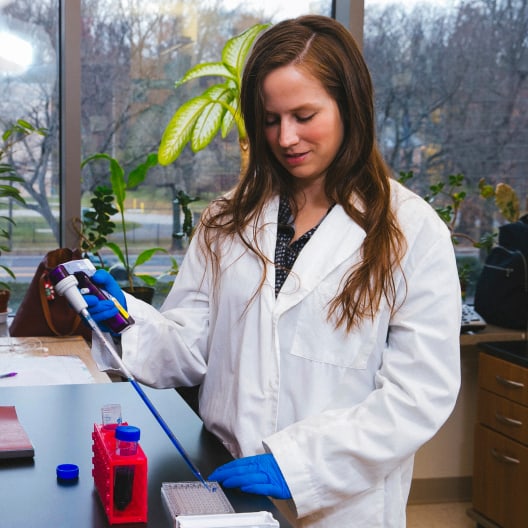 A woman wearing a white lab coat and blue gloves uses a pipette in a laboratory setting, working with a tray of test tubes and scientific equipment on the counter. Large windows and plants are in the background.