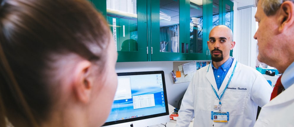 A man in a white lab coat speaks to two colleagues in a laboratory. He stands by a computer, with green cabinets and lab equipment visible in the background.
