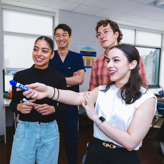 Four young adults stand together in a bright office, smiling and engaged. One woman in front holds a whiteboard marker and gestures enthusiastically, while the others watch attentively.