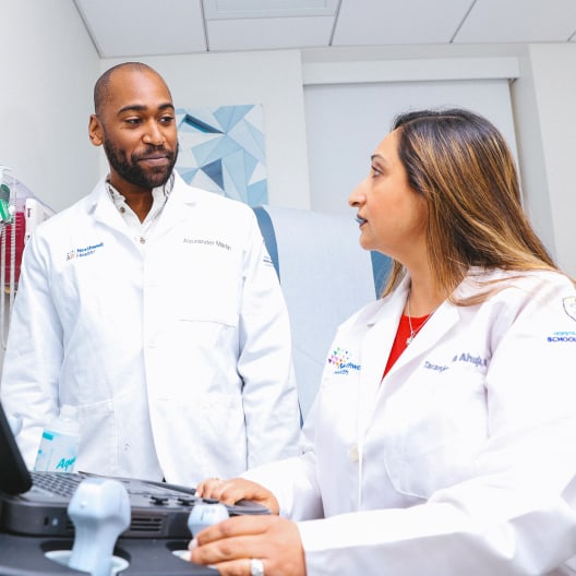Two healthcare professionals in white lab coats have a conversation in a medical office. One is standing and the other is seated at a computer, both appearing focused and engaged. Medical equipment is visible in the foreground.