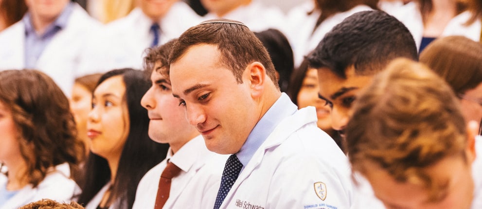 A group of medical students wearing white coats gathers closely together indoors, smiling and talking during what appears to be a professional or academic event.