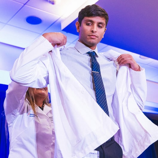A young man in business attire receives help putting on a white coat during a ceremony, symbolizing his entry into the medical profession. Blue lighting and a professional setting are visible in the background.