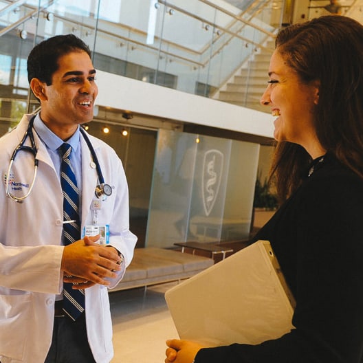 A doctor in a white coat and stethoscope smiles and talks with a woman holding a folder in a bright, modern building with glass railings and stairs in the background.