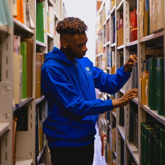 A person wearing a blue hoodie browses books on a shelf in a brightly lit library aisle, surrounded by colorful books.