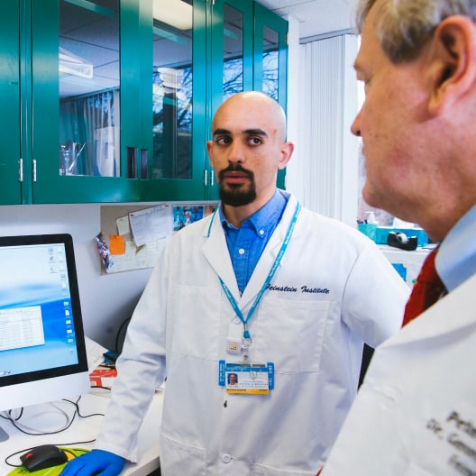 A man in a white lab coat speaks to two colleagues in a laboratory. He stands by a computer, with green cabinets and lab equipment visible in the background.