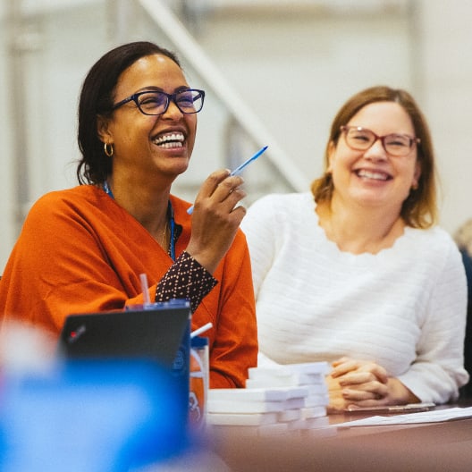 Two women are sitting at a table, smiling and laughing. One woman wears glasses and an orange top, holding a pen, while the other wears glasses and a white sweater. Papers and a laptop are on the table in front of them.