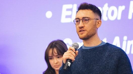 A man in glasses speaks into a microphone in front of a purple screen with white text, while a woman stands slightly behind him.