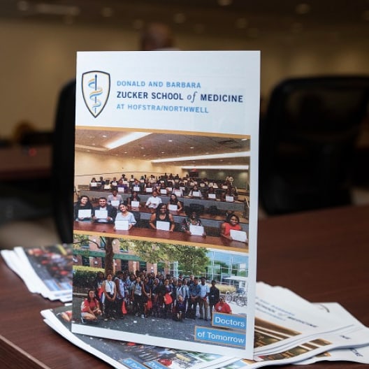 A brochure for the Zucker School of Medicine at Hofstra/Northwell is displayed on a table, featuring group photos of students and the text Doctors of Tomorrow.