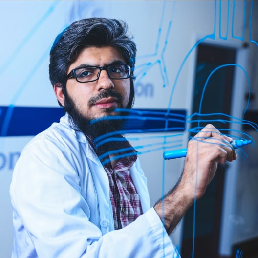 A man wearing glasses and a white lab coat writes with a blue marker on a clear board, with blue lines and shapes visible on the surface in front of him.