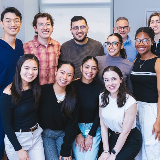 A diverse group of eleven people, standing closely together and smiling at the camera in a brightly lit indoor setting, suggesting a friendly and positive atmosphere.