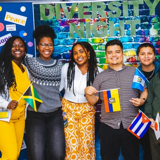 Five smiling people stand together holding different national flags in front of a colorful brick wall with the words Diversity Night above them. The atmosphere is joyful and celebratory.