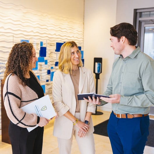 Three people stand indoors, smiling and conversing. One woman holds a folder, another stands with hands clasped, and a man holds an open notebook. They appear to be in a professional or office setting.