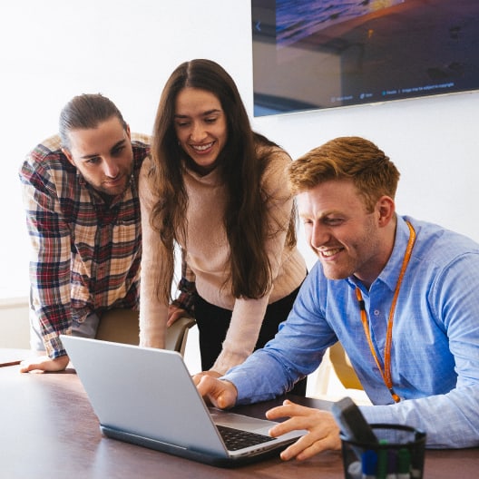 Three people gather around a laptop in an office, smiling and looking at the screen, appearing engaged and collaborative. A large monitor is visible on the wall in the background.