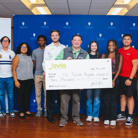 A group of eight people stand smiling in front of a blue backdrop, holding an oversized check for $3,000 made out to The Pipeline Program from Jovia.