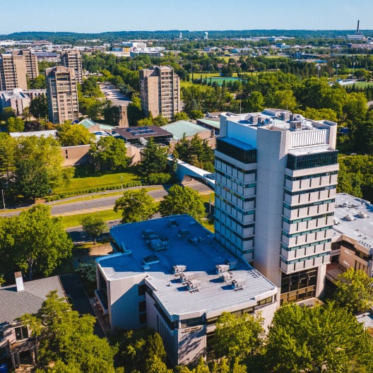 Aerial view of a modern multi-story building surrounded by trees, with other buildings, green spaces, and a distant cityscape in the background on a clear day.