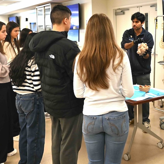 A group of students stands around a table while a man holds an anatomical model, giving a demonstration in a classroom or medical lab setting.