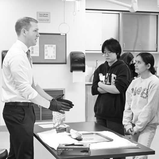 A man wearing gloves speaks to two students standing by a table with medical supplies in a classroom or lab setting.