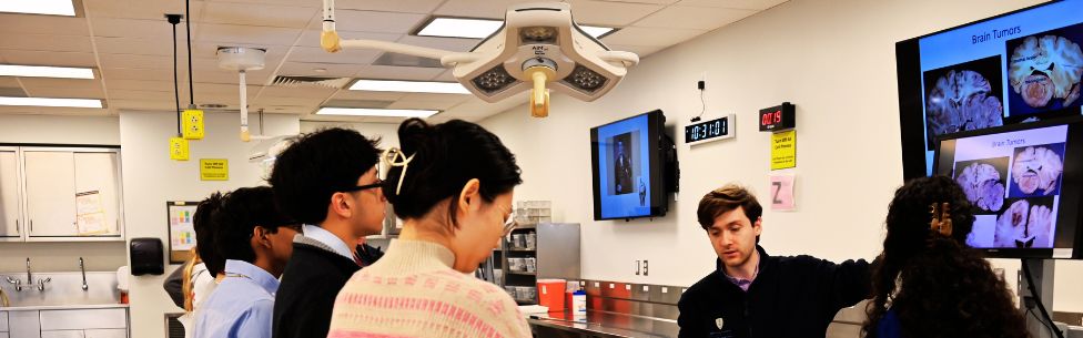 A group of students listens to an instructor presenting brain images on large screens in a brightly lit lab classroom with medical equipment.