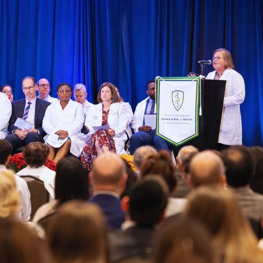 group of faculty on stage wearing white coat. one doctor at podium speaking